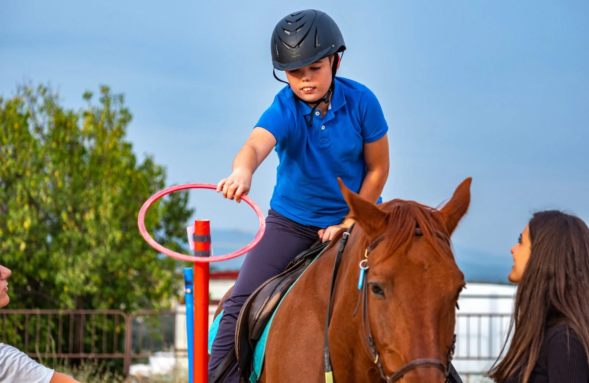 Un niño a caballo coloca un aro en un poste acompañado por su terapeuta