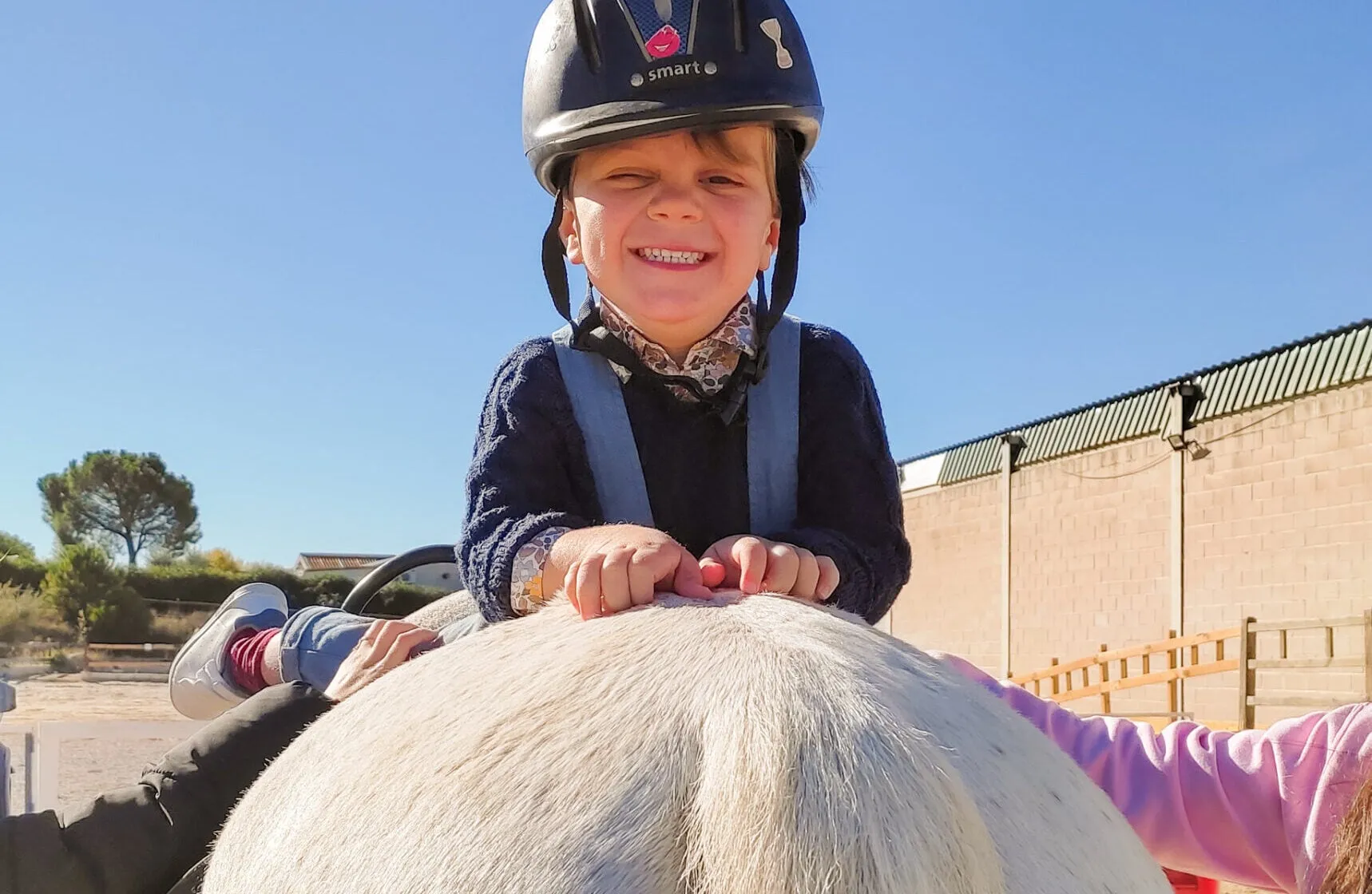 Un niño con casco apoya las manos en la grupa de un caballo blanco