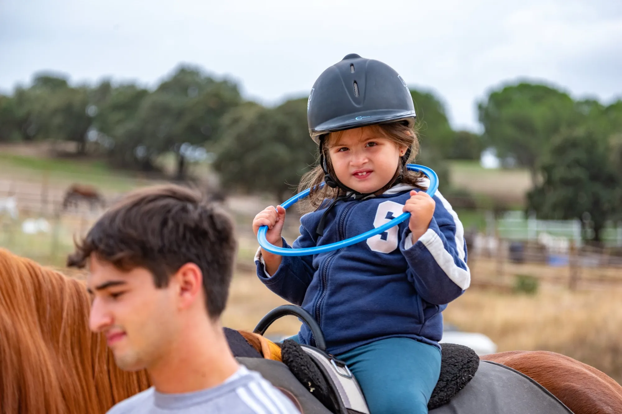 Una niña con casco juega con un aro subida al caballo junto a un voluntario