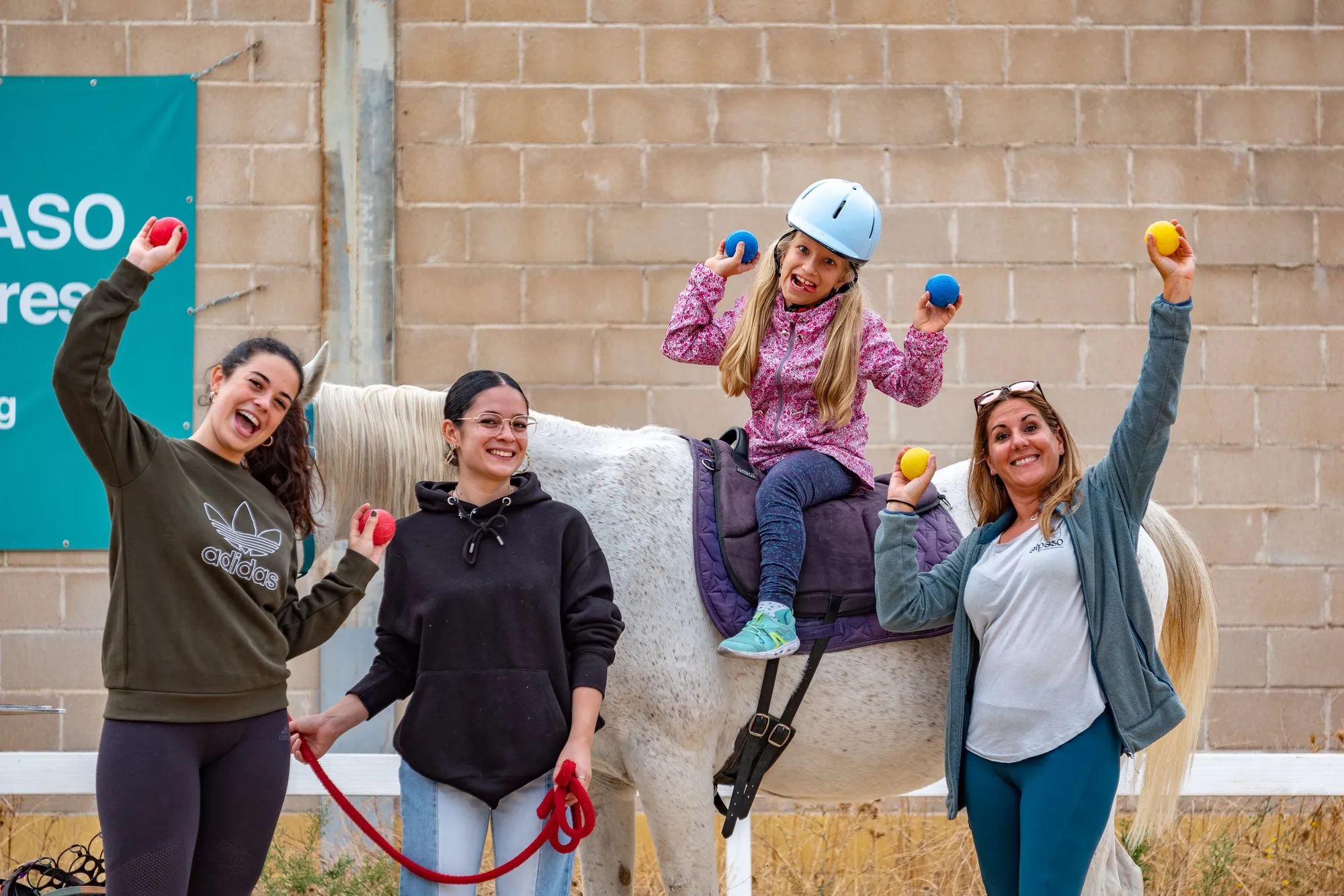 Una niña a caballo celebra con tres terapeutas levantando pelotas de colores